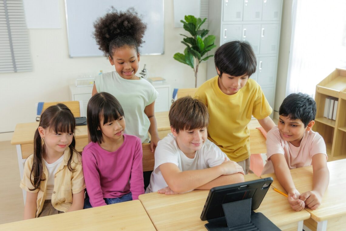 A group of diverse elementary school students are gathered around a tablet