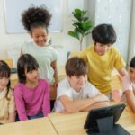 A group of diverse elementary school students are gathered around a tablet